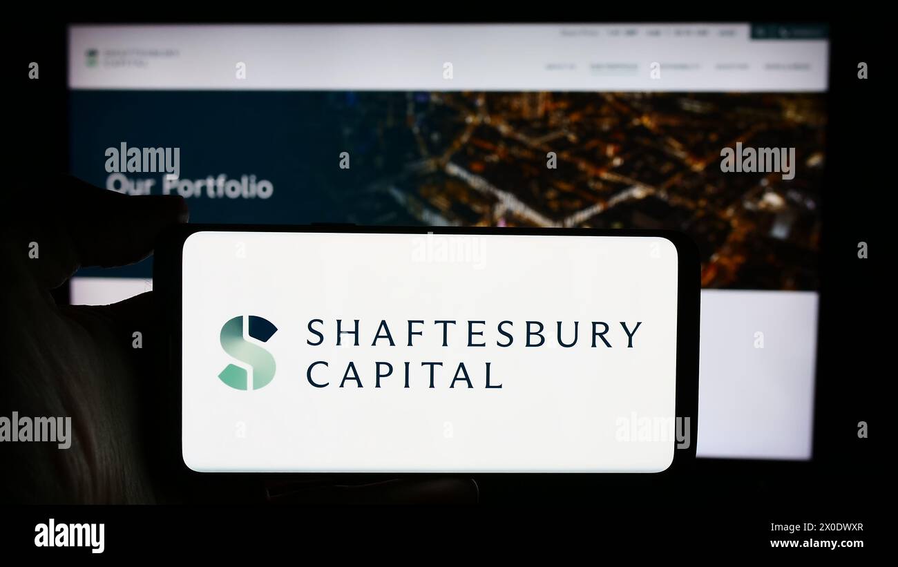 Person, die ein Mobiltelefon mit dem Logo der britischen Investmentgesellschaft Shaftesbury Capital plc vor der Webseite hält. Schwerpunkt auf dem Display des Telefons. Stockfoto