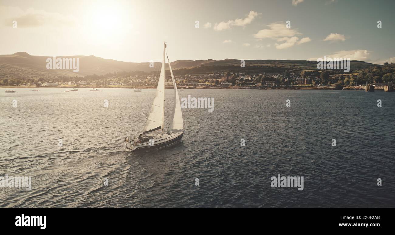 Blick aus der Luft auf das Sonnenlicht auf dem Segelboot bei Reflexion der Meeresbucht. Hochlandseeküste mit Wald und Bergen bei hellem Sonnenlicht. Im Hafen von Brodick gibt es keine Naturlandschaften mit Luxusyachten, die im Kino gedreht werden Stockfoto