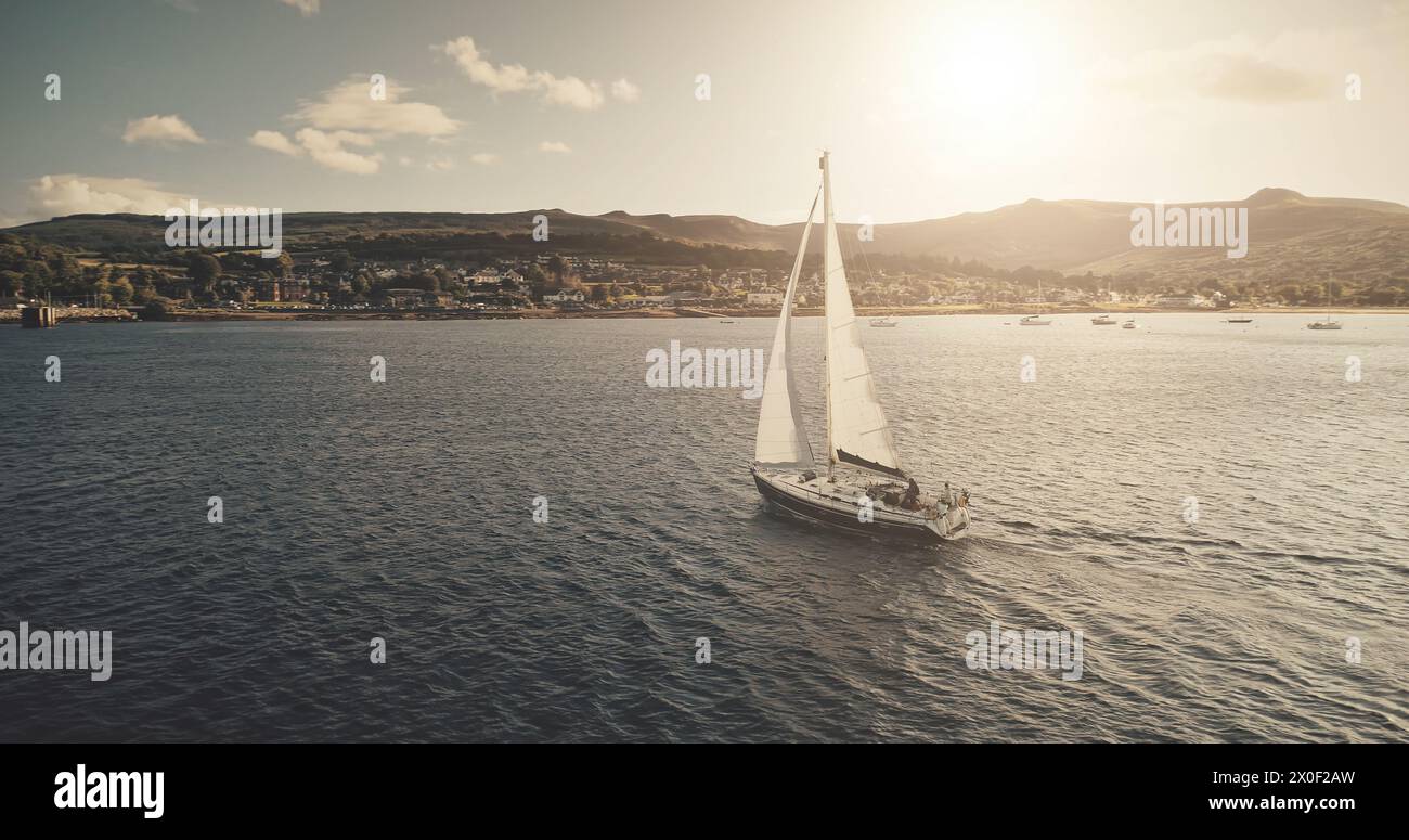 Blick aus der Luft auf das Sonnenlicht auf dem Segelboot bei Reflexion der Meeresbucht. Hochlandseeküste mit Wald und Bergen bei hellem Sonnenlicht. Im Hafen von Brodick gibt es keine Naturlandschaften mit Luxusyachten, die im Kino gedreht werden Stockfoto