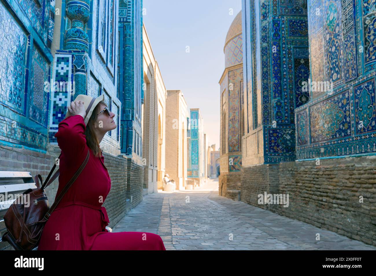 Frau mit Hut und rotem Kleid, die sich auf der Bank entspannt und die Schönheit des alten Shah-i-Zinda-Komplexes in Samarkand, Usbekistan, genießt Stockfoto