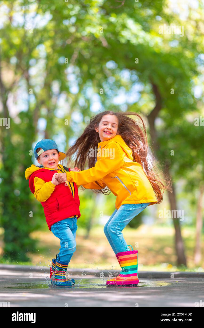 Glückliche Kinder Mädchen und Jungen in bunten Gummi-Regenstiefeln spielen draußen und springen in regnerischer Pfütze Stockfoto