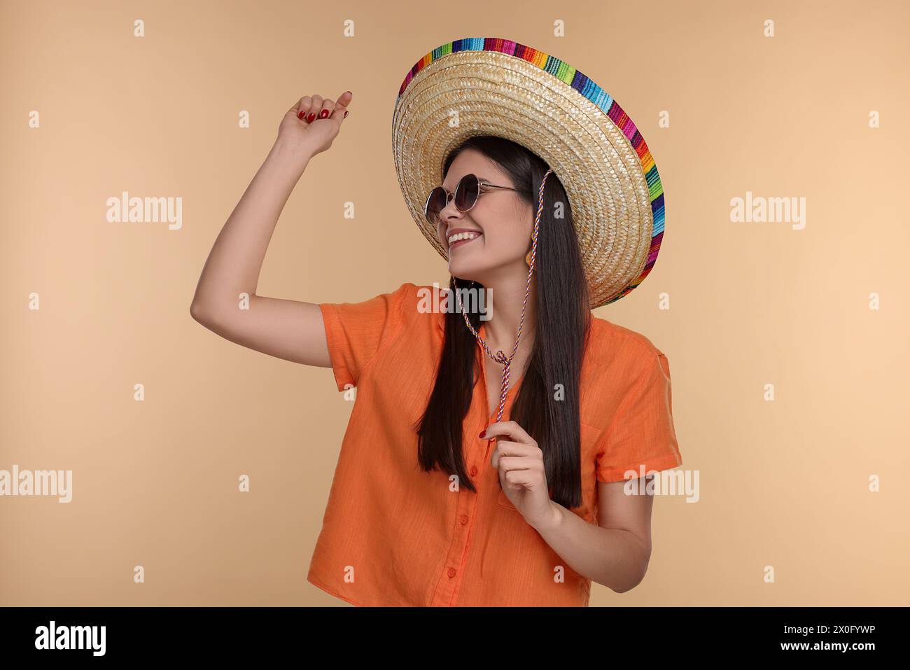 Junge Frau mit mexikanischem Sombrero-Hut und Sonnenbrille, die auf beigefarbenem Hintergrund tanzt Stockfoto