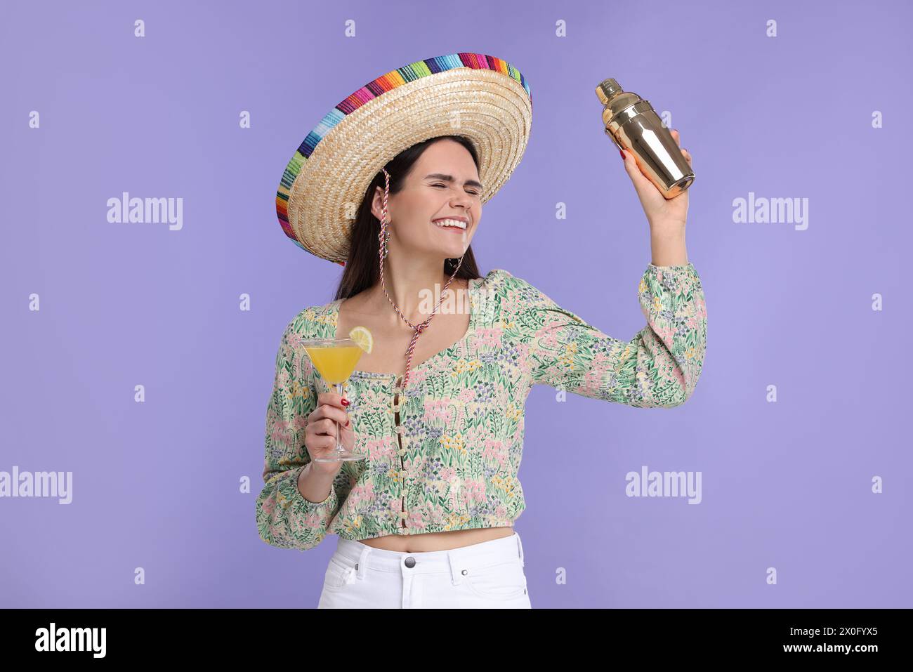 Junge Frau in mexikanischem Sombrero-Hut mit Cocktail und Shaker auf violettem Hintergrund Stockfoto