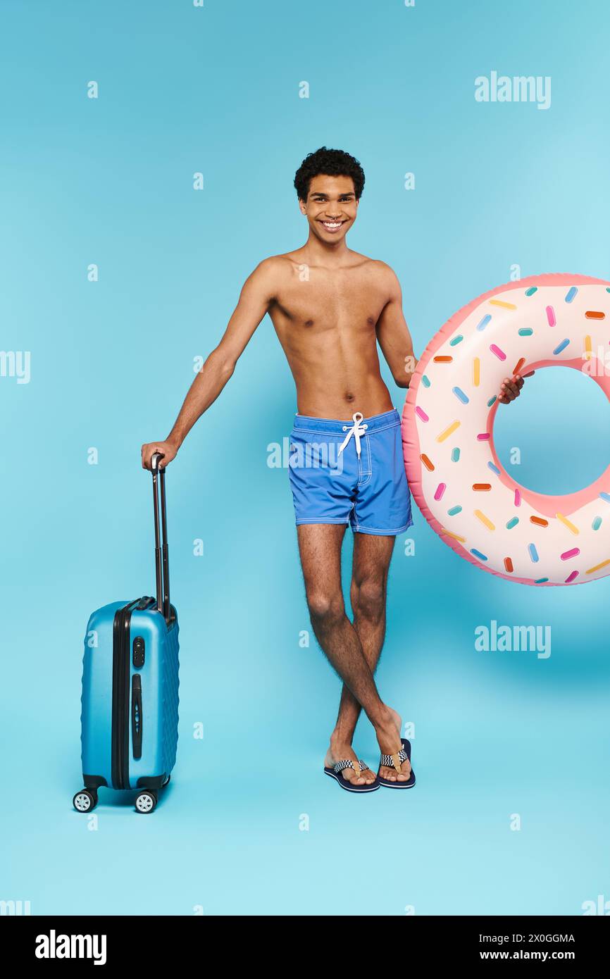 Fröhlicher afroamerikanischer Mann in Badehosen mit Schwimmring und Koffer, der vor der Kamera lächelt Stockfoto