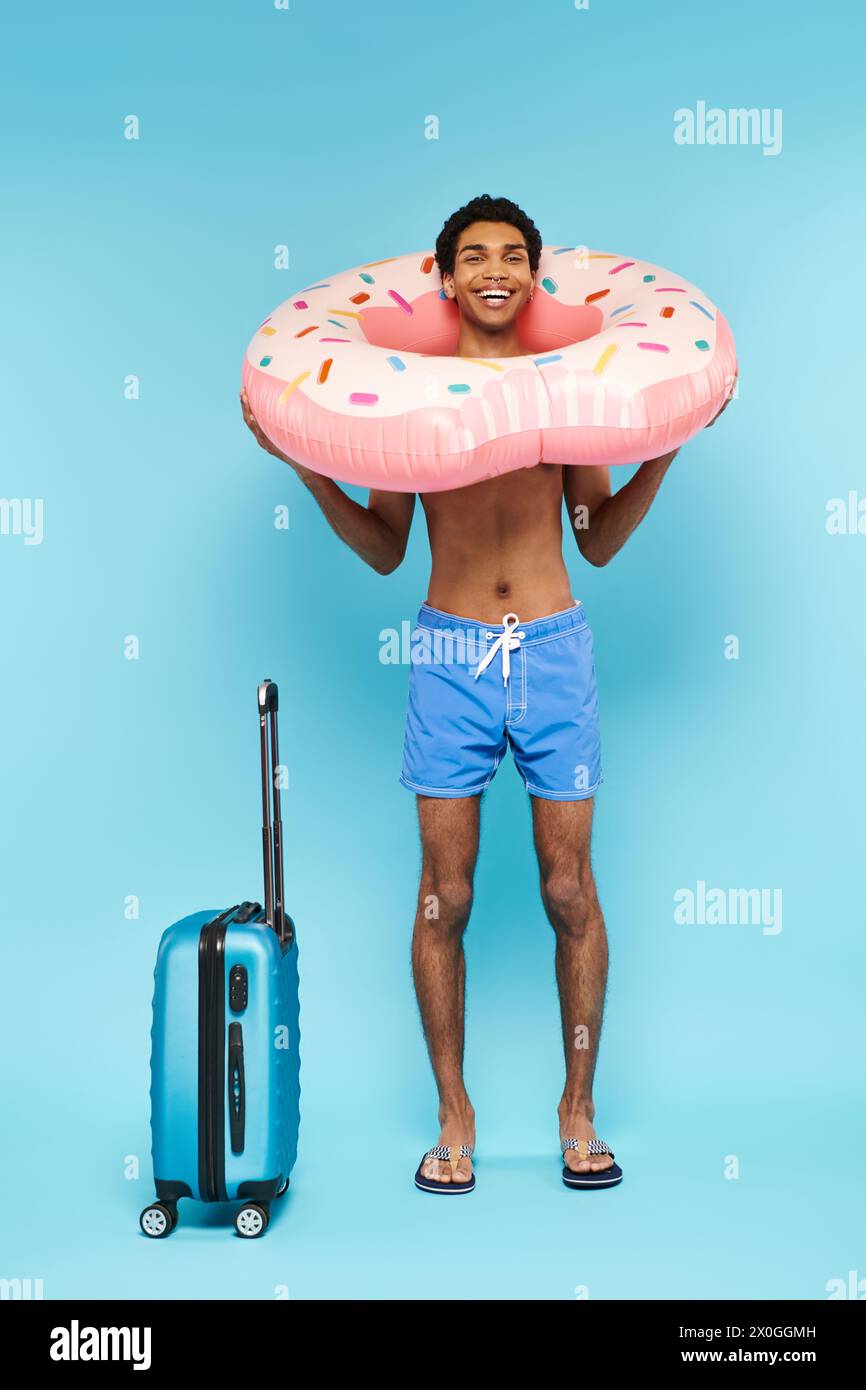 fröhlicher afroamerikanischer Mann in Badehosen mit Schwimmring und Koffer, der vor der Kamera lächelt Stockfoto