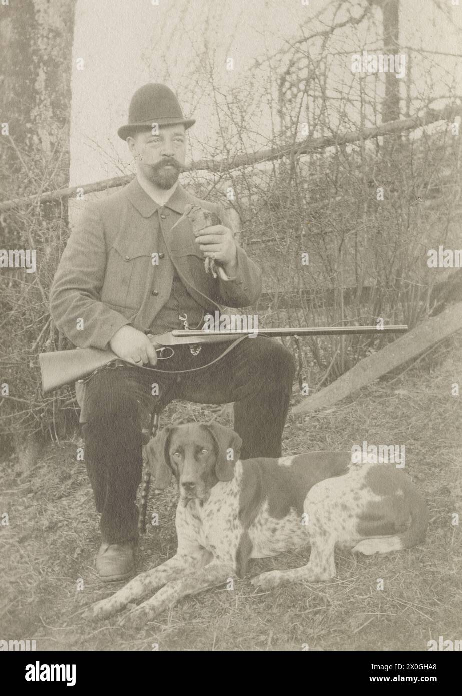 Hunter, etwa 40 Jahre alt, trägt einen dreiteiligen dunklen Anzug mit steifem Kragen und Krawatte, sitzt auf einem Felsen mit auseinander liegenden Beinen. Er hält eine Doppelflinte in der Hand. Er trägt einen getrimmten Vollbart und eine Nickelbrille. Er hat seinen Hut jämmerlich zur Seite geschoben. Vor ihm, im hohen Gras, liegt sein Jagdhund, wahrscheinlich ein deutscher Hund. Junge Nadelbäume im Hintergrund [automatisierte Übersetzung] Stockfoto