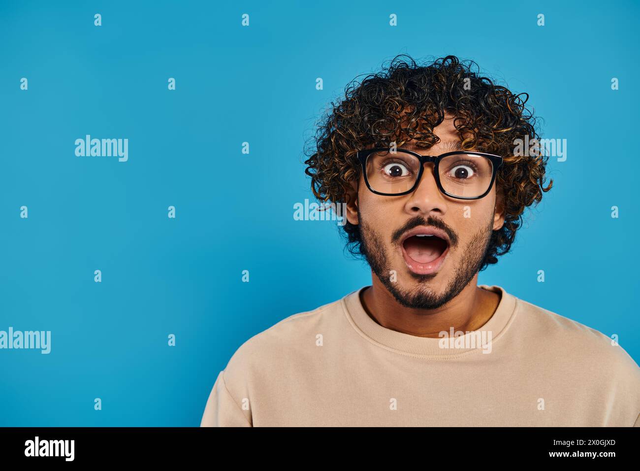 Ein indischer Student mit lockigem Haar und Brille sieht vor blauem Hintergrund überrascht aus. Stockfoto