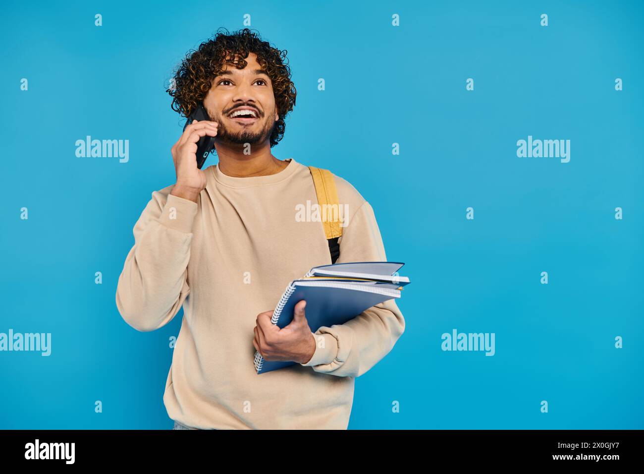 Ein indischer Student in lässiger Kleidung steht vor blauem Hintergrund, hält eine Mappe und spricht mit einem Handy. Stockfoto