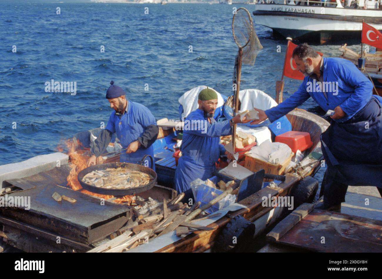 Zwei Männer stehen auf einem Boot am Goldenen Horn in Istanbul in einer großen Pfanne und braten Fisch. Einer der Männer übergibt einem dritten Mann auf dem Steg etwas. Die Männer tragen blaue Overalls. [Automatisierte Übersetzung] Stockfoto