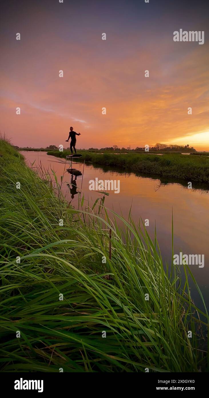 Tragflächenreiter gleitet mit seinem Brett in einem der Kanäle der Ria de Aveiro in Portugal während des Sonnenuntergangs über dem Wasser. Stockfoto