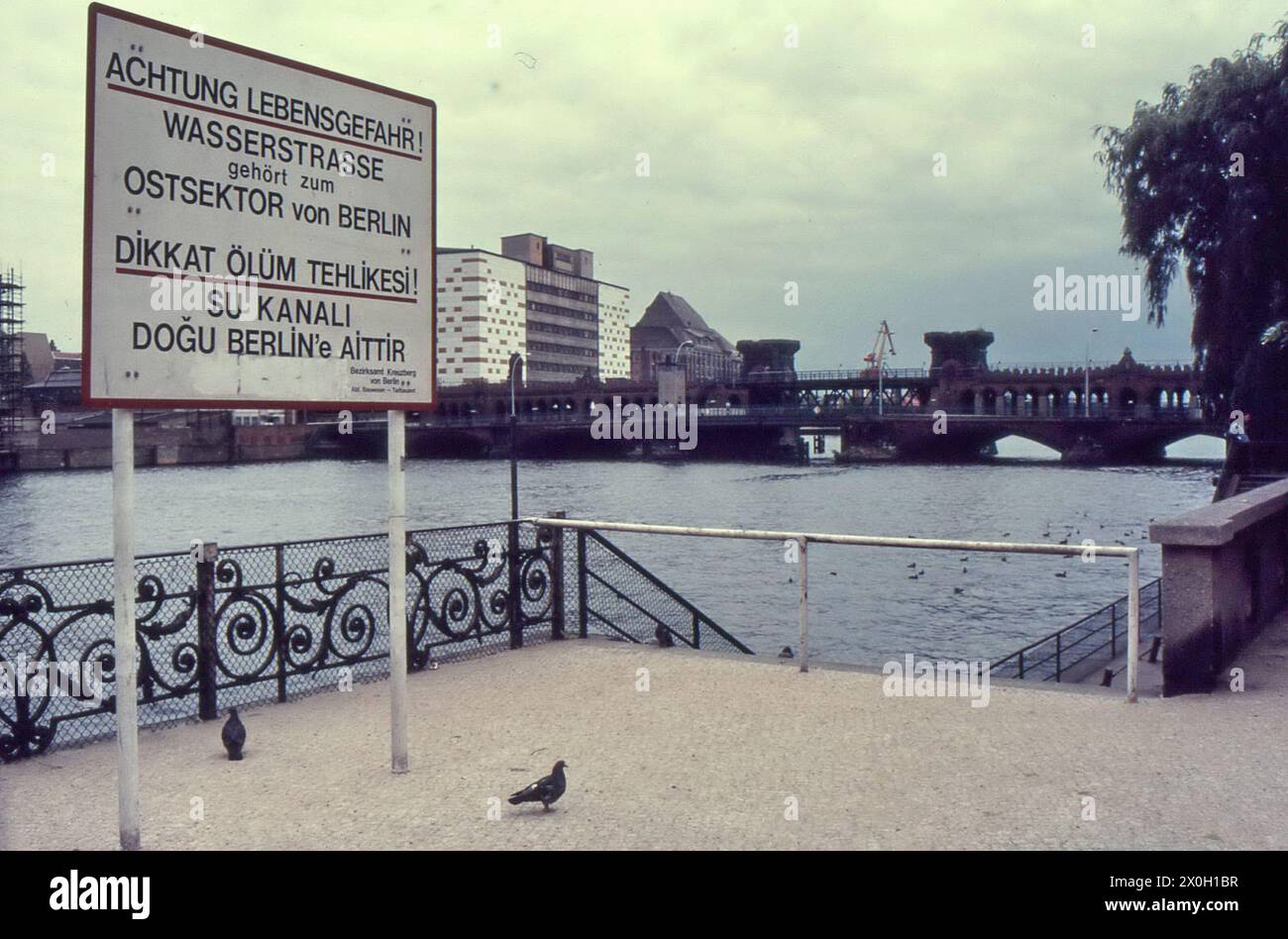 Ein Schild mit der Aufschrift "Vorsicht Gefahr! Die Wasserstraße gehört zum östlichen Teil Berlins. Im Hintergrund befindet sich die Oberbaumbrücke. Stockfoto