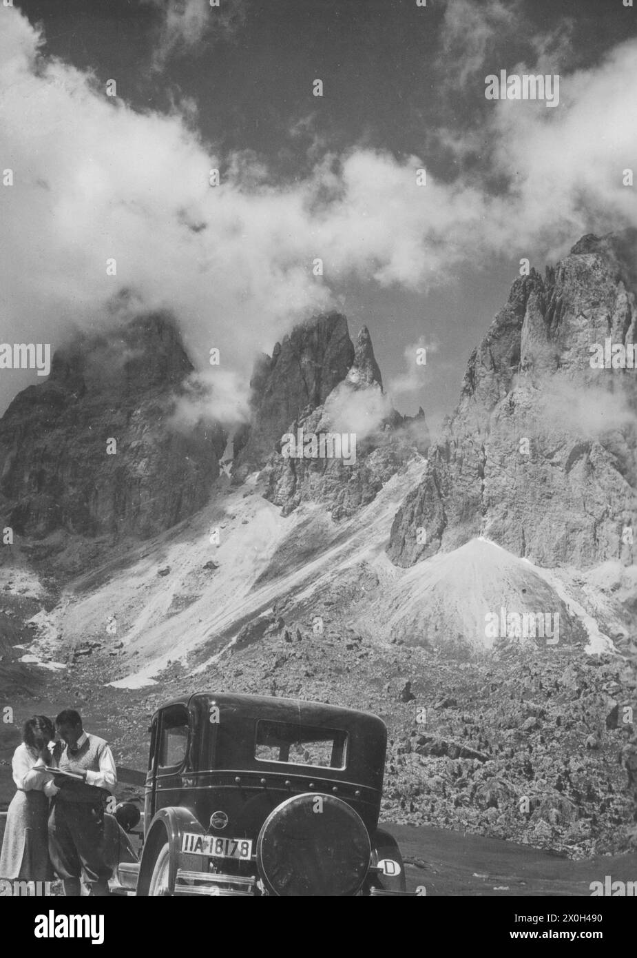 Ein Lesepaar steht neben einer Huppmobile Serie E in den italienischen Dolden mit einem Bergmassiv im Hintergrund. Meine Eltern sagten mir, dass sie manchmal nur die steileren Teile der Pässe rückwärts beherrschen könnten. Straßen waren noch nicht asphaltiert und es war besser mit dem Rückwärtsgang [automatisierte Übersetzung] Stockfoto