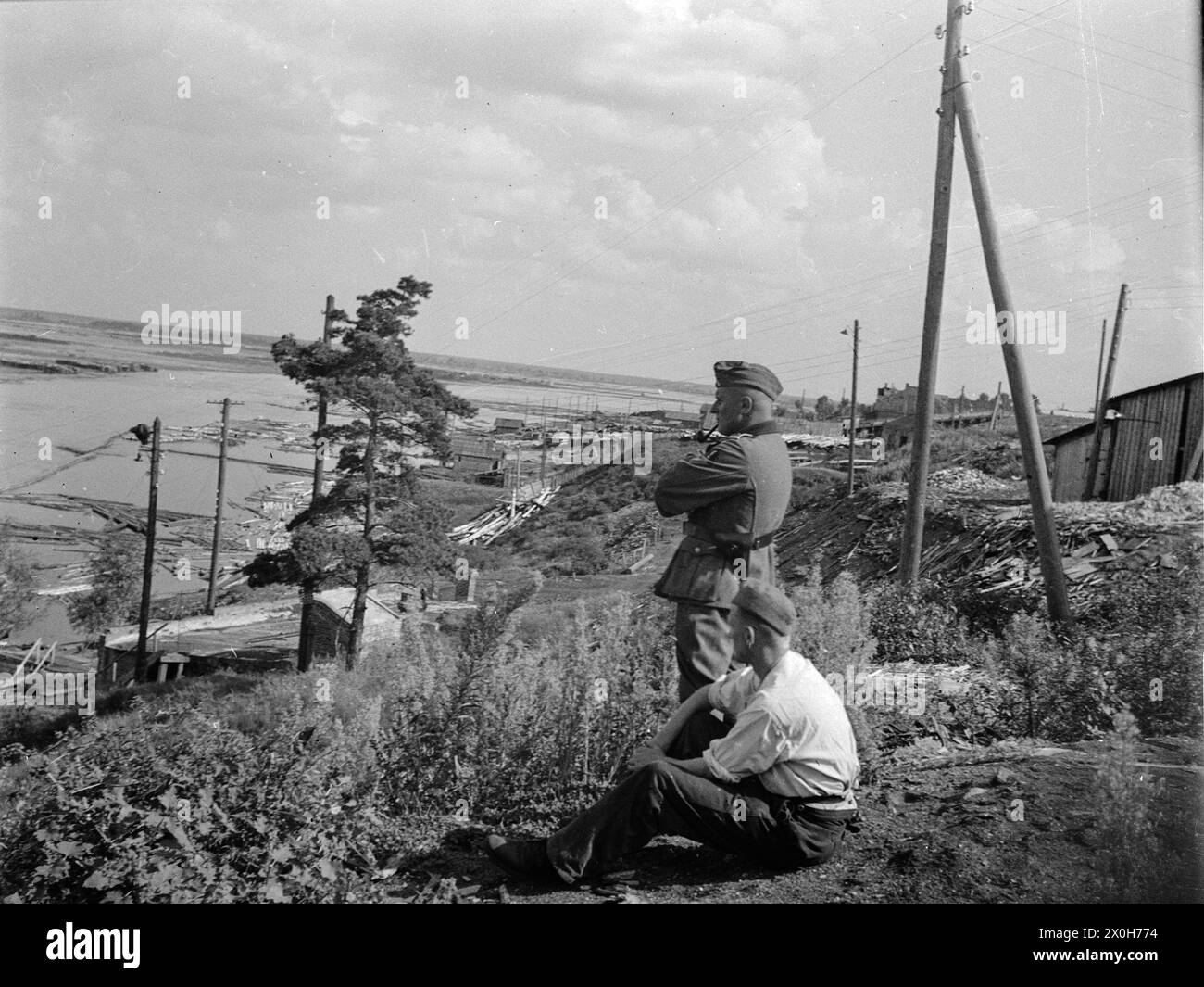 Unzählige Baumstämme werden in einem hölzernen Hafen am Dnieper gesammelt. Zwei deutsche Soldaten sitzen im Gras. Das Bild wurde von einem Mitglied des Radfahrgrenadierregiments 2/Radfahrsicherungsregiment 2 im Nordteil der Ostfront aufgenommen. [Automatisierte Übersetzung] Stockfoto