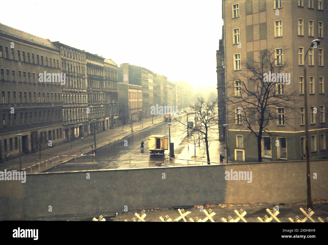 Ein Schulausflug nach Berlin war damals üblich. Für mich war es auch eine Reise in die Gedächtnisspur. Der Blick schweift über einen Bahnsteig in der Bernauerstraße in eine Seitenstraße. Die abgebildete Mauer ist bereits die zweite Mauer, die äußere Mauer, und zeigt Ost-Berlin. Sie können das sehen, weil die Panzerabsperrungen zwischen den beiden Wänden platziert wurden. Herzzerreißende Szenen fanden hier nach dem 13. August 1961 statt. Um Platz für die Grenzbefestigung und den Todesstreifen zu schaffen und Fluchtwege zu verhindern, wurde die gesamte Ost-Berliner Fassade abgerissen. Die Schwefeldämpfe sind spürbar, je weiter man in den Osten blickt Stockfoto