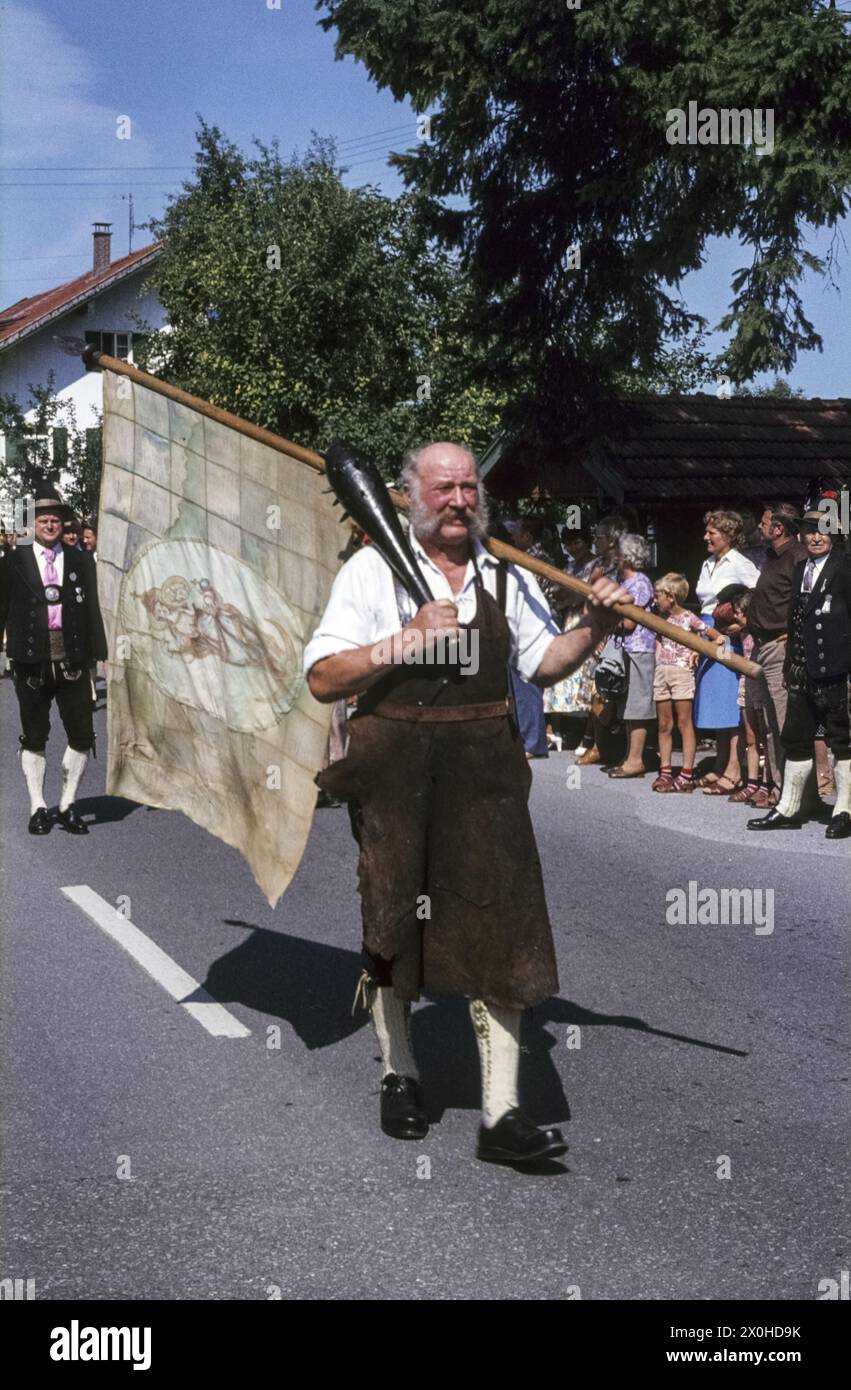 Ein Mann stellt den Schmied von Kochel bei der jährlichen Kostümparade unter Mariä Himmelfahrt dar. Er trägt eine Lederschürze und eine Fahne. [Automatisierte Übersetzung] Stockfoto
