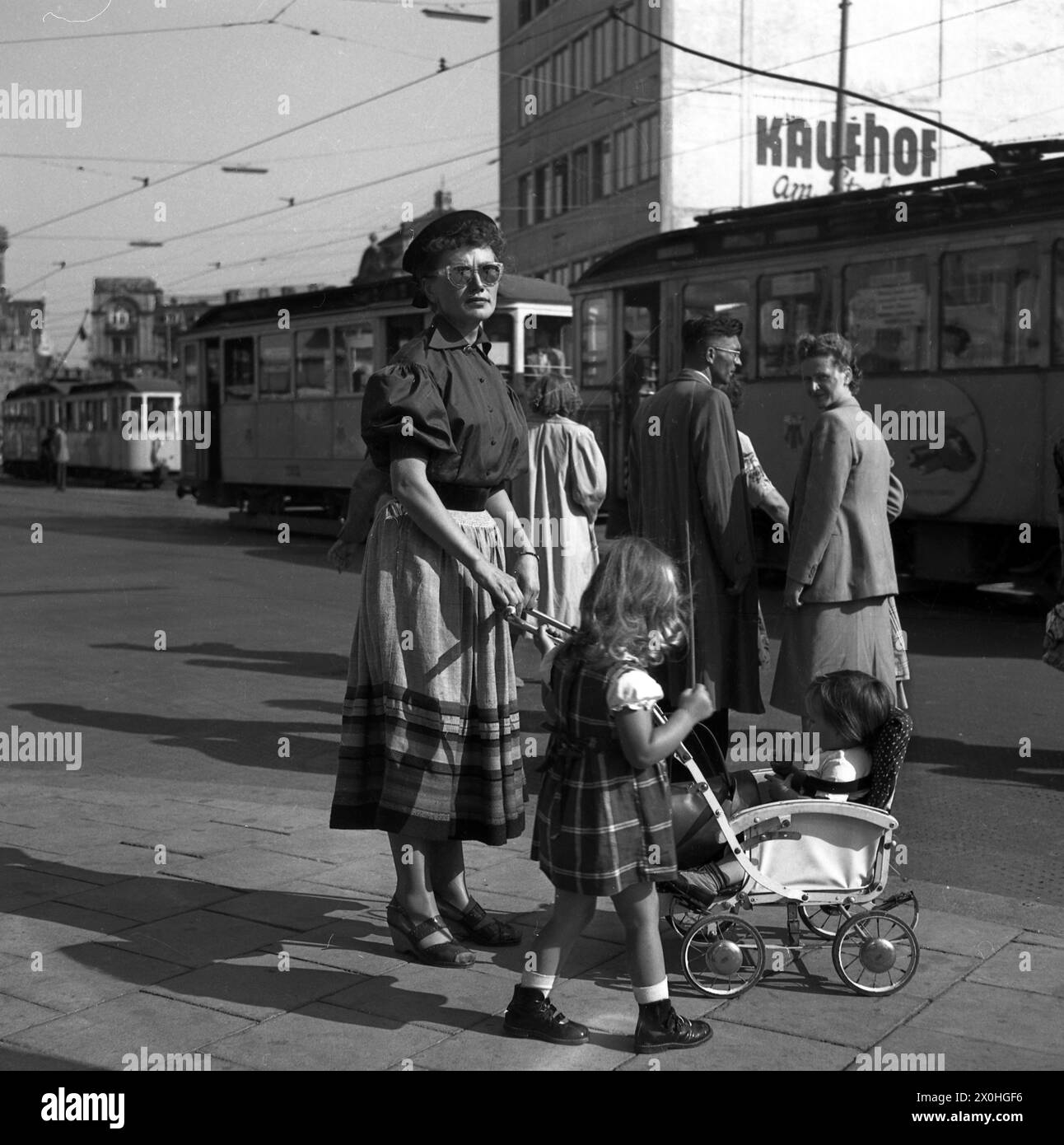 Eine Mutter mit Sonnenbrille steht mit einem Mädchen und einem Kleinkind in einem Kinderwagen vor einer Straßenbahn in einer Großstadt. [Automatisierte Übersetzung] Stockfoto