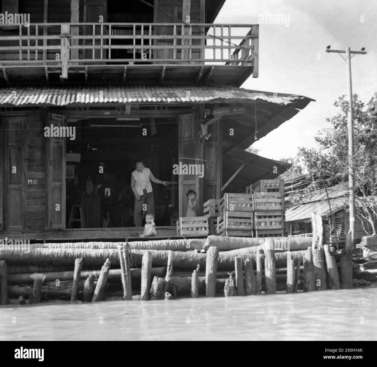 Ein Wohnhaus am Fluss Chao Phraya in Bangkok, Thailand. Vor dem Haus sind zwei kleine Kinder. Im Hauseingang steht eine alte Frau. Das Haus liegt direkt am Flussufer. Die Bank ist mit Baumstämmen befestigt. Vor dem Haus befinden sich einige Holzkisten. [Automatisierte Übersetzung] Stockfoto