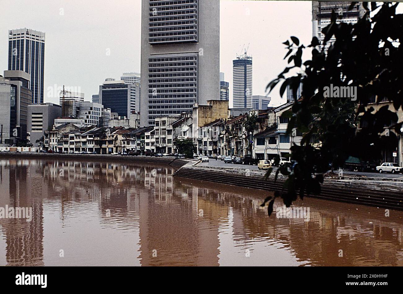 Blick entlang einer Strandpromenade in Singapur. Im Vordergrund rotbraunes Wasser. An der Bank eine Straße. Dahinter alte Häuser mit heruntergekommenen Fassaden und Dächern. Im Hintergrund neue Bürohochhäuser. [Automatisierte Übersetzung] Stockfoto