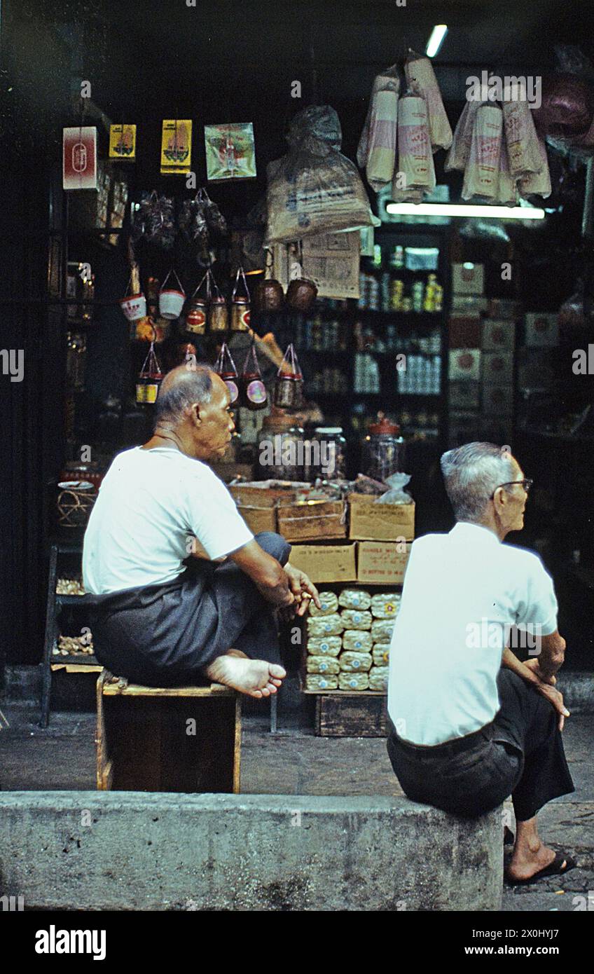 Zwei ältere Herren in hellen Hemden und dunklen Hosen sitzen auf dem Bürgersteig vor einem Geschäft in Singapur. Der Mann links im Bild sitzt auf einem Holzkasten oder einer Art Hocker, der Mann rechts im Bild sitzt auf einem Betonvorsprung. Im Hintergrund ist der Laden sichtbar, zahlreiche Waren hängen von der Decke, liegen in Kisten oder stehen auf Regalen. [Automatisierte Übersetzung] Stockfoto