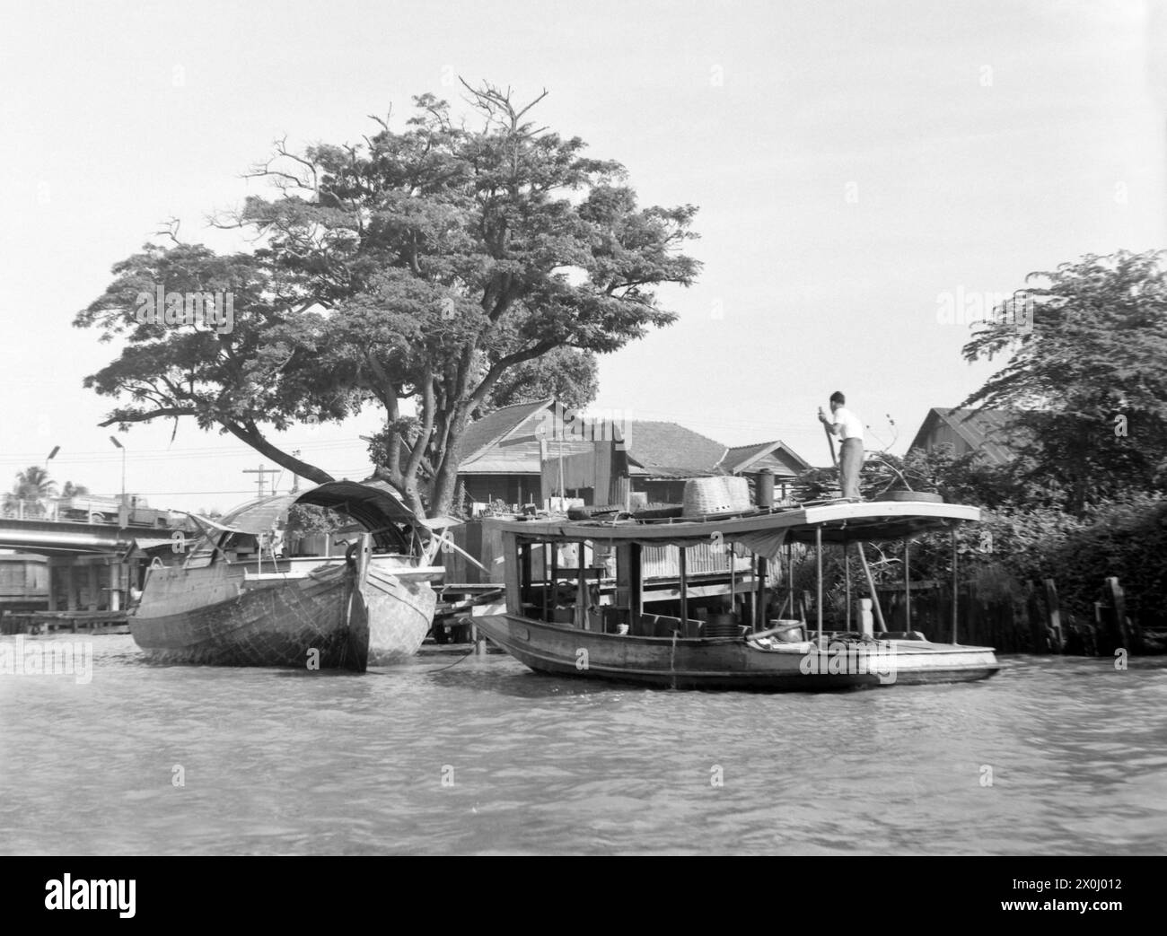 Blick auf den Fluss Chao Phraya in Bangkok im Juni 1972 in Thailand. Zwei große Boote sind auf dem Fluss. Auf dem Hinterboot ist ein Mann mit langem Pfosten. Hinter den Booten sieht man die Bank mit einigen Häusern, Hütten und Bäumen. [Automatisierte Übersetzung] Stockfoto