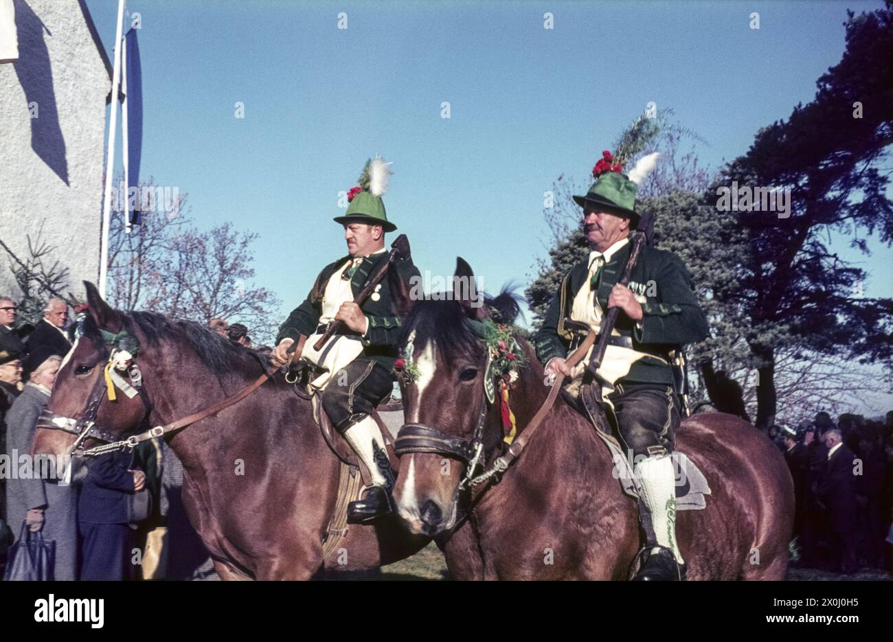 Am 6. November fahren zwei Reiter in Tracht mit Hüten und Schlümpfen auf dem Tölzer Leonhardi. Im Hintergrund sieht man Zuschauer. Undatiertes Foto, Mitte der 1960er Jahre. [Automatisierte Übersetzung] Stockfoto