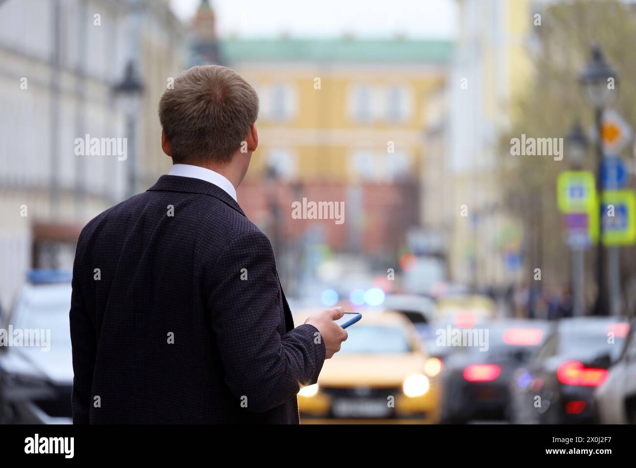 Mann in einem Business-Anzug, der mit Smartphone auf einer Straße auf Autohintergrund steht. Mit dem Mobiltelefon in der Stadt Stockfoto