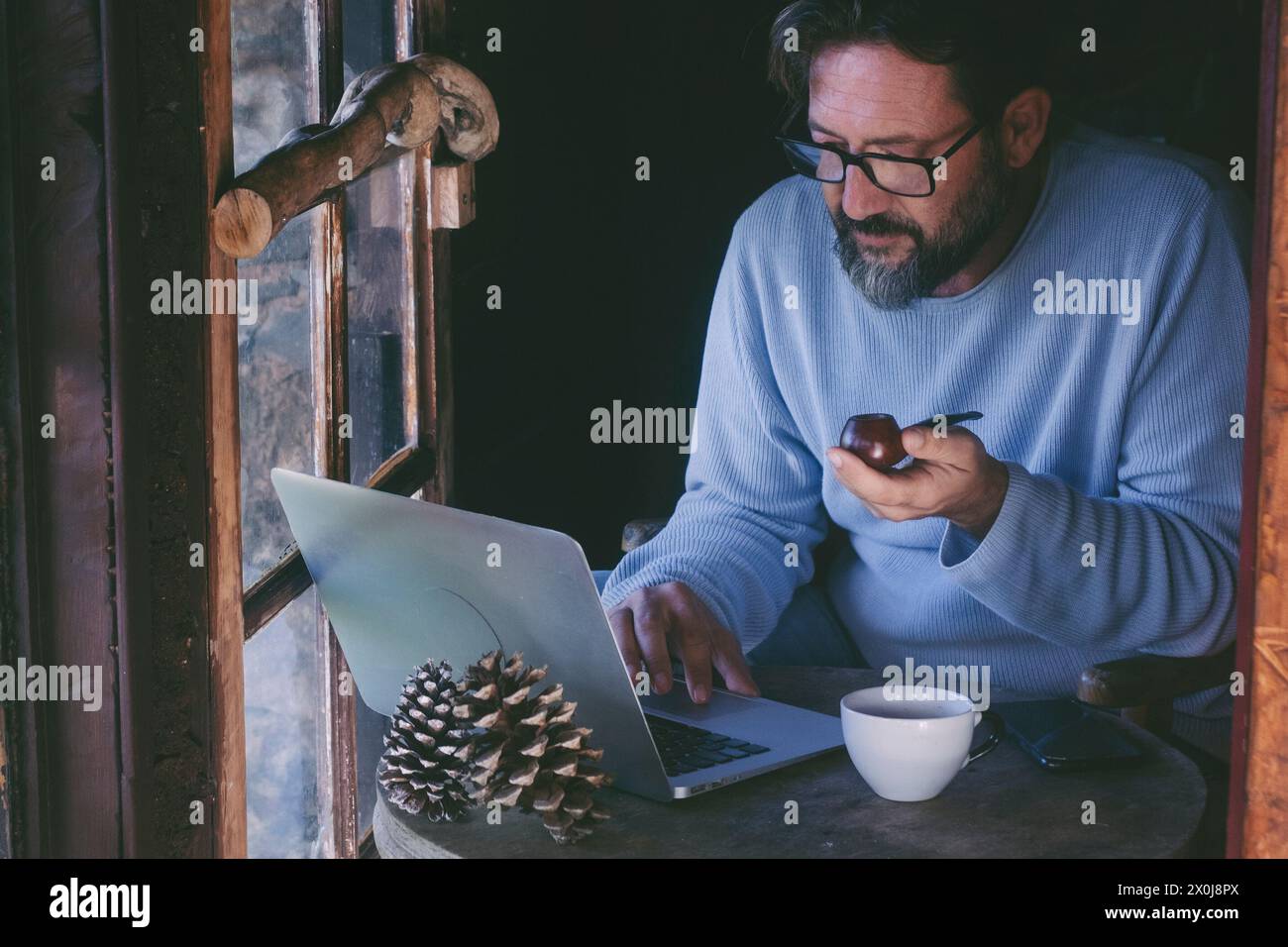 Ein reifer Mann sitzt zu Hause und arbeitet an einem Laptop-Computer und raucht Pfeife. Alternative Single Lifestyle männliche Leute innen in gemütlichem Hütte Holzchalet. Telearbeit online Stockfoto
