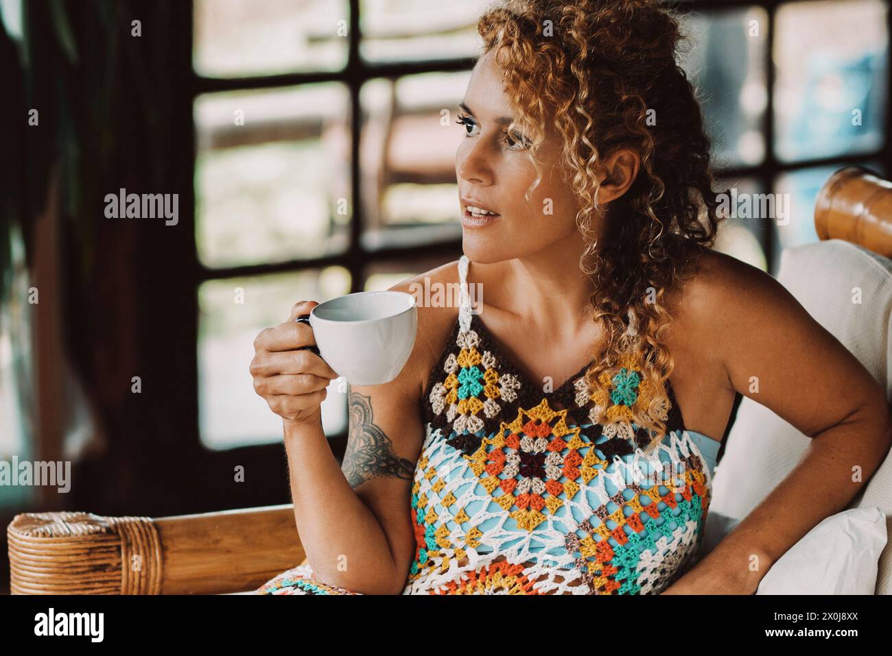 Elegante attraktive junge Erwachsene Frau mit warmem, wollfarbenem Strickkleid zu Hause beim Trinken aus dem Becher und beim Blick nach draußen - Konzept von trendigen schönen weiblichen Menschen, die Freizeit genießen, entspannen Sie sich drinnen auf dem Sofa Stockfoto