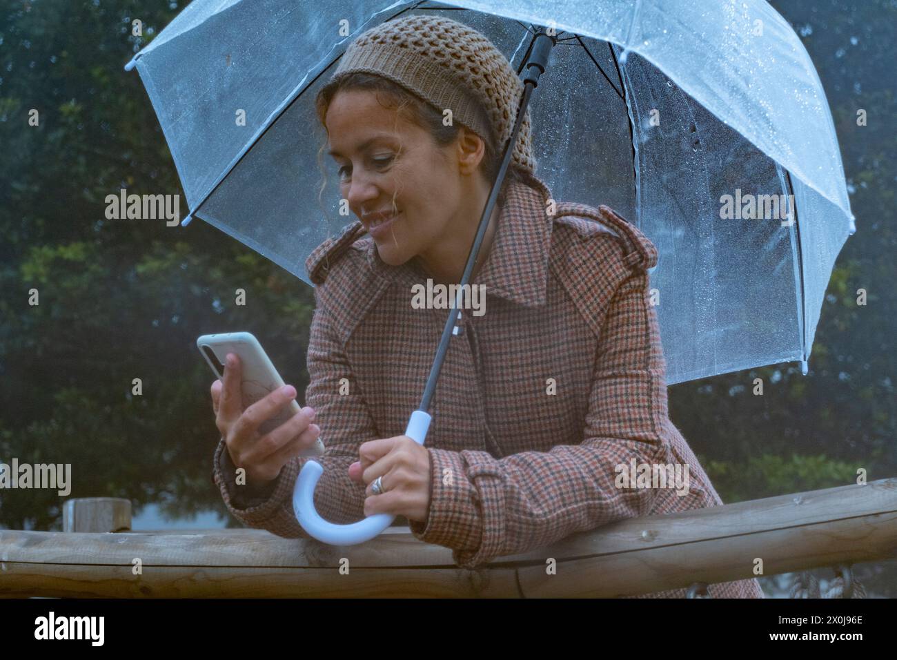 Eine Frau, die im Freien unter dem Regen mit einem transparenten Regenschirm telefoniert. Moderne Frau in der Technologie Freizeitbeschäftigung allein chatten und Nachrichten auf zellulärem Gerät senden. Menschen sind verbunden Stockfoto