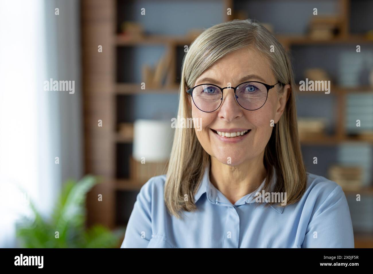 Porträt einer lächelnden Seniorin mit Brille, in blauer Bluse, in einer modernen Büroumgebung. Dieses Bild spiegelt ihr Selbstvertrauen und ihre Professionalität wider. Stockfoto