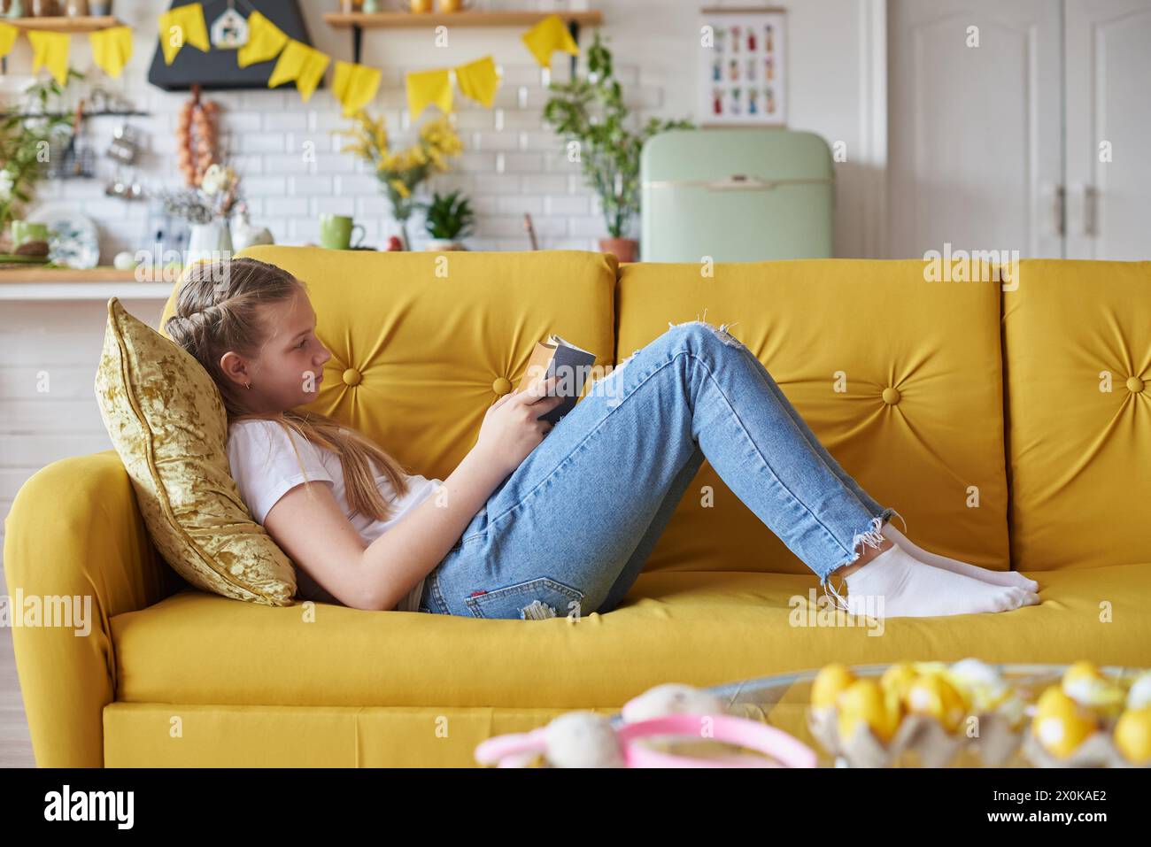 Mädchen, 12 Jahre alt, liegt auf dem Sofa im Wohnzimmer und liest ein Buch. Stockfoto