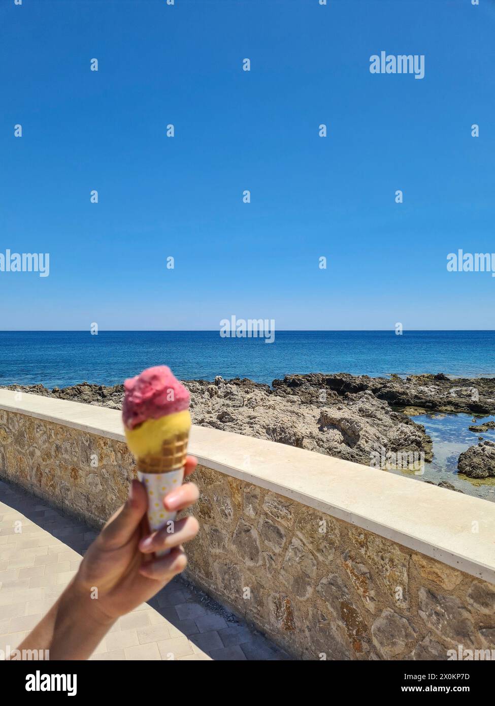 Die Hand eines kleinen Kindes im Vordergrund hält zwei Kugeln Eis in einem Kegel, an der Promenade von Cala Ratjada, blauer Himmel am Horizont während der Sommerferien auf Mallorca, Spanien Stockfoto