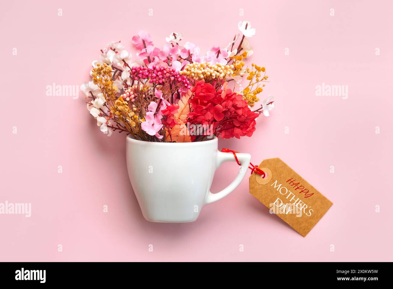 Alles Gute zum Muttertag. Weiße Tasse Kaffee mit verschiedenen Blumen und braunem Verkaufsanhänger mit dem Text Happy Mothers Day auf rosa Hintergrund Stockfoto