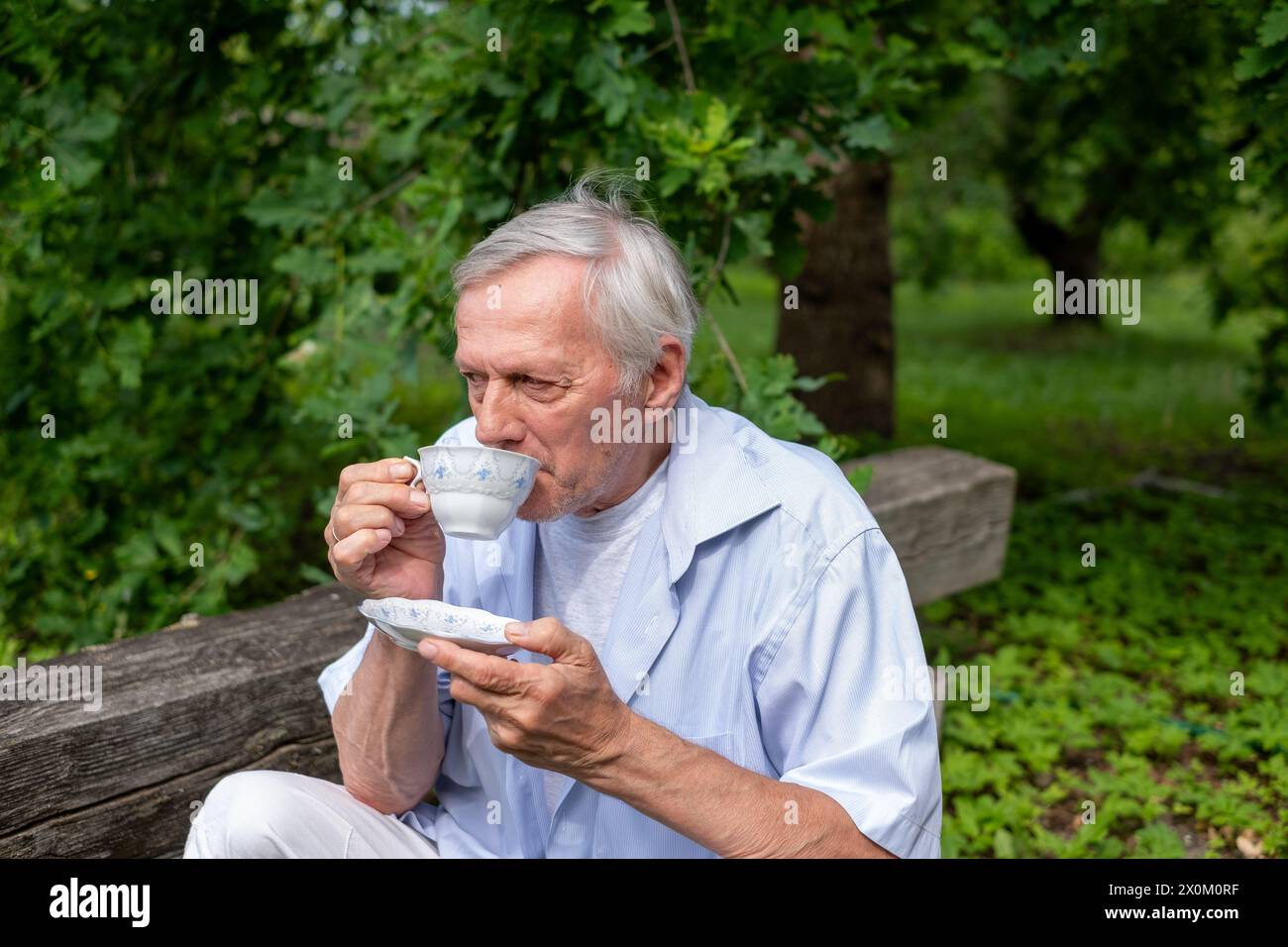 Seniorenmann, der auf einer Holzbank mit Tee ruht, inmitten von üppigem Grün, der Inbegriff eines ruhigen ländlichen Lebens. Hochwertige Fotos Stockfoto