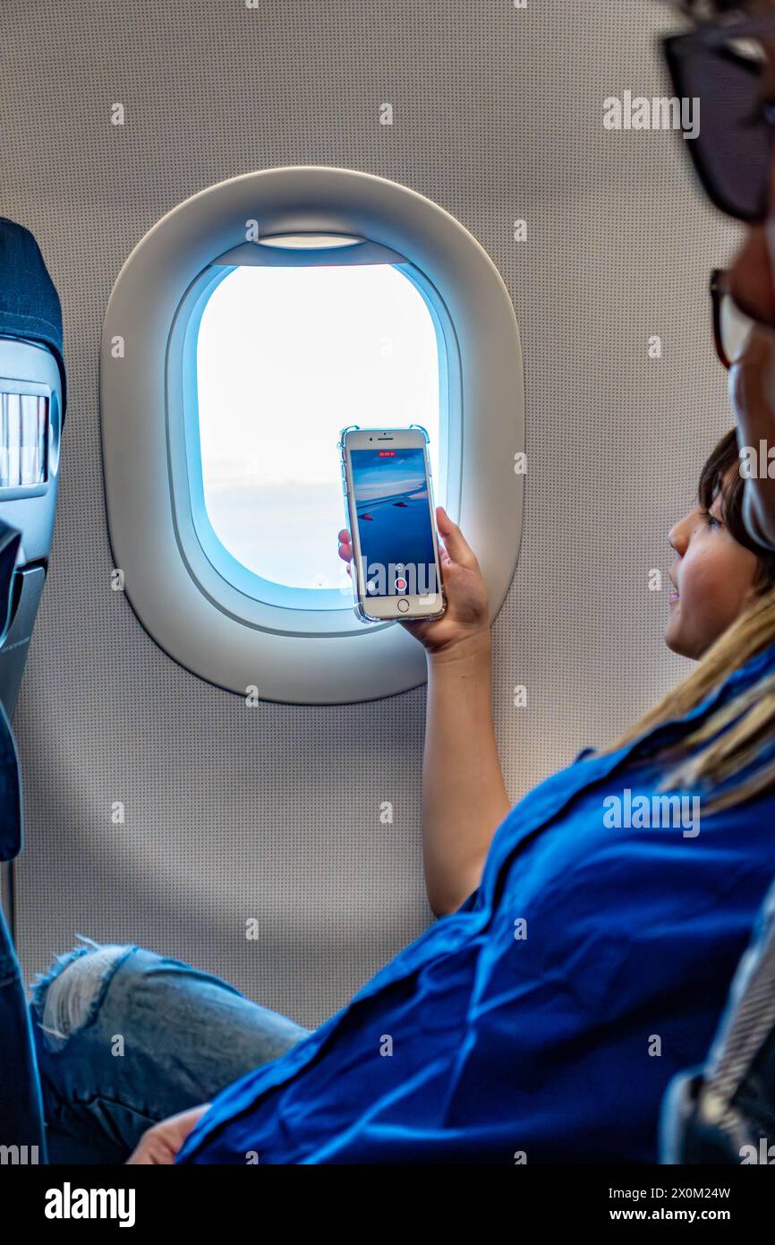 Ein Junge nimmt den Blick aus einem Fenster eines Flugzeugs auf seinem Handy auf. Stockfoto