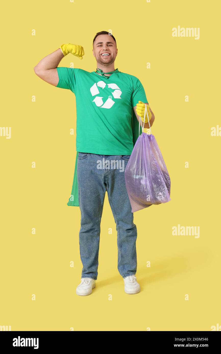 Männlicher Freiwilliger im superheldencape mit Müllsack, der Muskeln auf gelbem Hintergrund zeigt. Abfallrecyclingkonzept Stockfoto