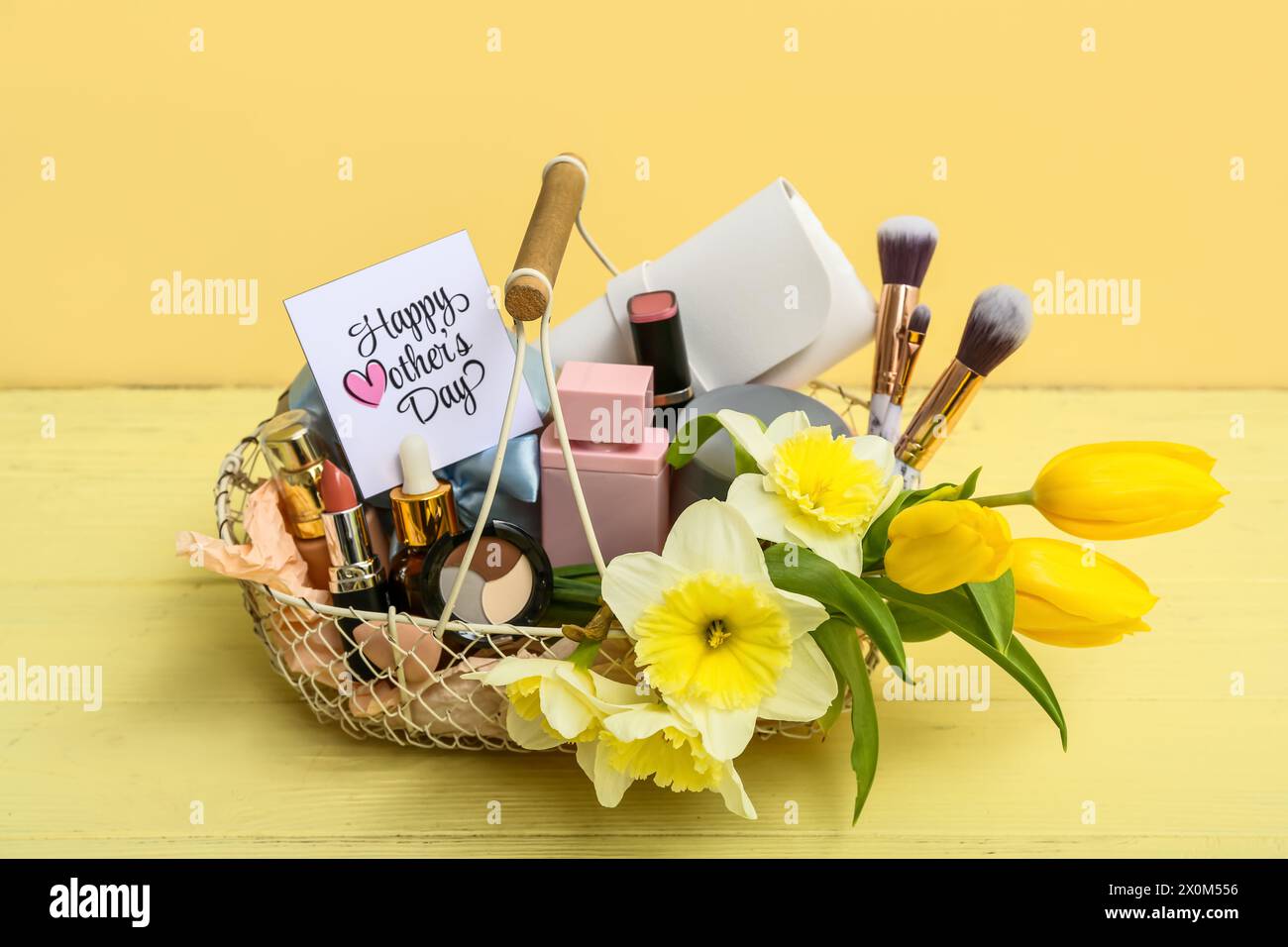 Geschenkkorb mit dekorativen Kosmetikartikeln, Make-up-Bürsten und Narzissen-Blumen für die Muttertagsfeier auf farbigem Holztisch Stockfoto