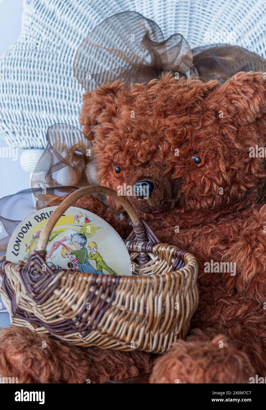 Der alte Teddybär hält einen antiken Eierkorb mit einem Kinderrekord aus den 1940er Jahren, in dem Kinder spielen. Der Pelzbär sitzt auf einem alten Rattanstuhl. Display für Spielzeug. Stockfoto
