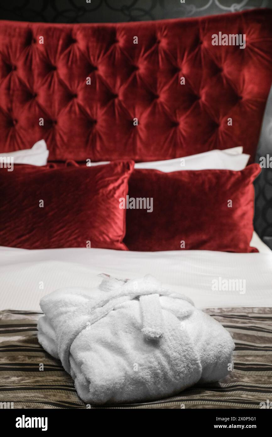 Gefalteter weißer Bademantel auf dem Bett vor dem Hintergrund von Kissen und Kissen rot gepolsterte Wand. Klassisches, luxuriöses Zimmer mit Innendesign für einen Schlaf Stockfoto