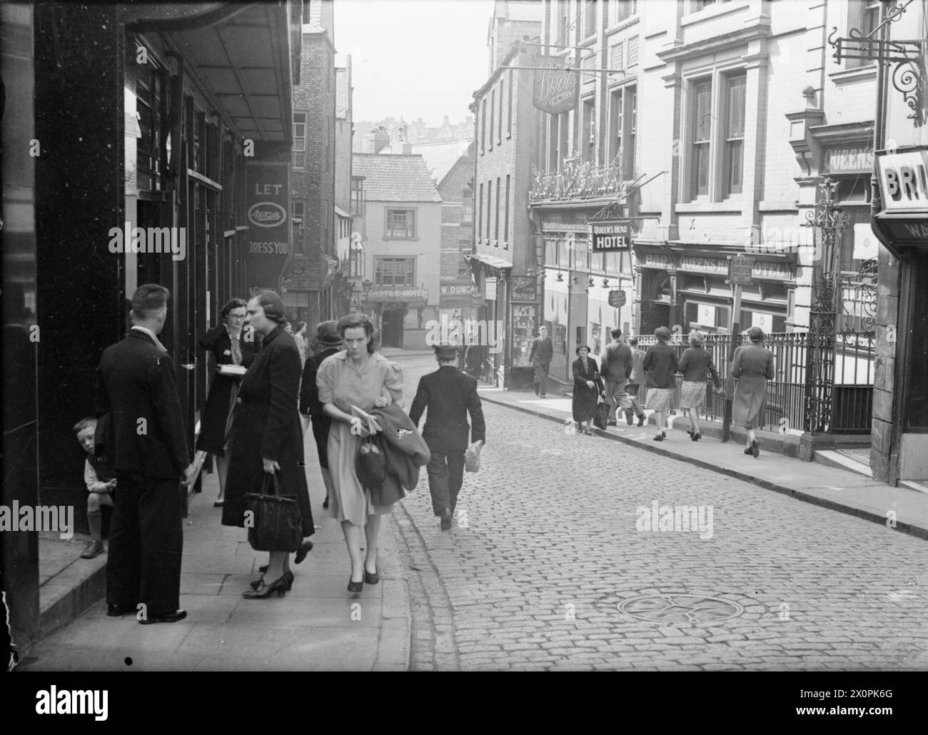 Ein NAHER OSTEN-SOLDAT BESUCHT BRITAIN: LIFE IN WARTIME DURHAM, ENGLAND, UK, 1943 - Eine geschäftige Szene, in der Käufer ihre täglichen Geschäfte in der Silver Street in Durham machen. Links ist das Bekleidungsgeschäft 'Burton' zu sehen. Im Hintergrund, am Ende der Straße, befindet sich das Castle Hotel und ein Geschäft namens „W Duncan“. Auf der rechten Seite befinden sich die Geschäfte Pullar's Dye Works und Boot's The Chemist. Das Queen's Head Hotel ist auch sichtbar, ebenso wie ein Schild für einen Luftschutzraum Stockfoto