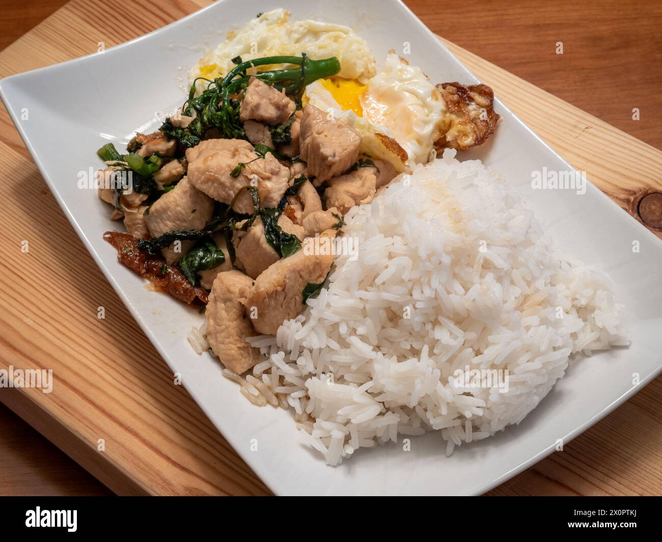 Genießen Sie die Aromen von thailändischem Basilikum-Huhn, serviert mit einem perfekt Spiegelei, Brokkolini und duftendem Jasminreis, elegant präsentiert auf einem weiß Stockfoto