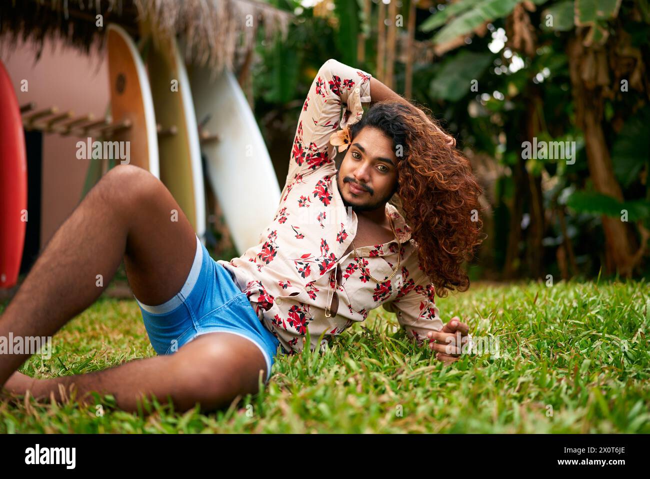 Selbstbewusster schwuler Mann mit kräftigen lockigen Haaren liegt im tropischen Garten, lebhafter Stil. Blaue Shorts und Hemd mit Blumenmuster sorgen für einen lebendigen Look. LGBT-Reisen, selbst Stockfoto