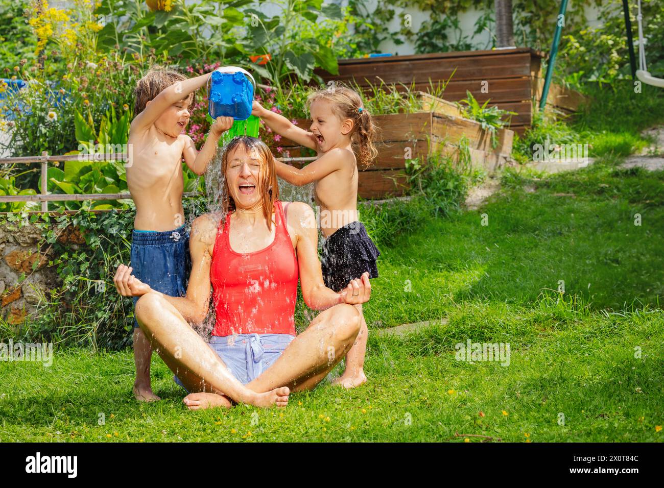 Zwei kleine Geschwister gießen spielerisch Wasser über ihre sitzende Mutter Stockfoto