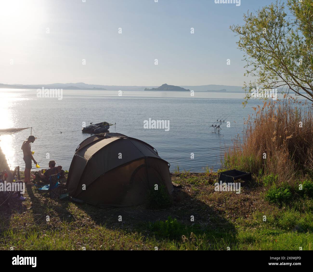 Zelt am Ufer des Bolsenasees mit Angelruten im Freien und einer Insel in der Ferne. Provinz Viterbo, Region Latium, Italien. April 2024 Stockfoto