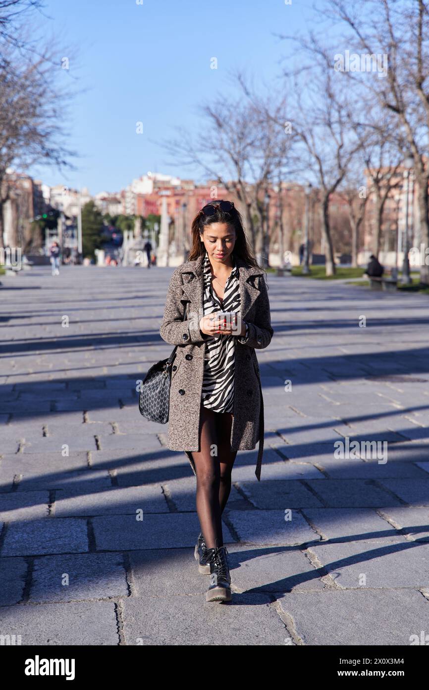 Vollständige Ansicht einer jungen Latina-Frau, die durch die Stadt läuft, mit ihrem Handy, um zu chatten Stockfoto