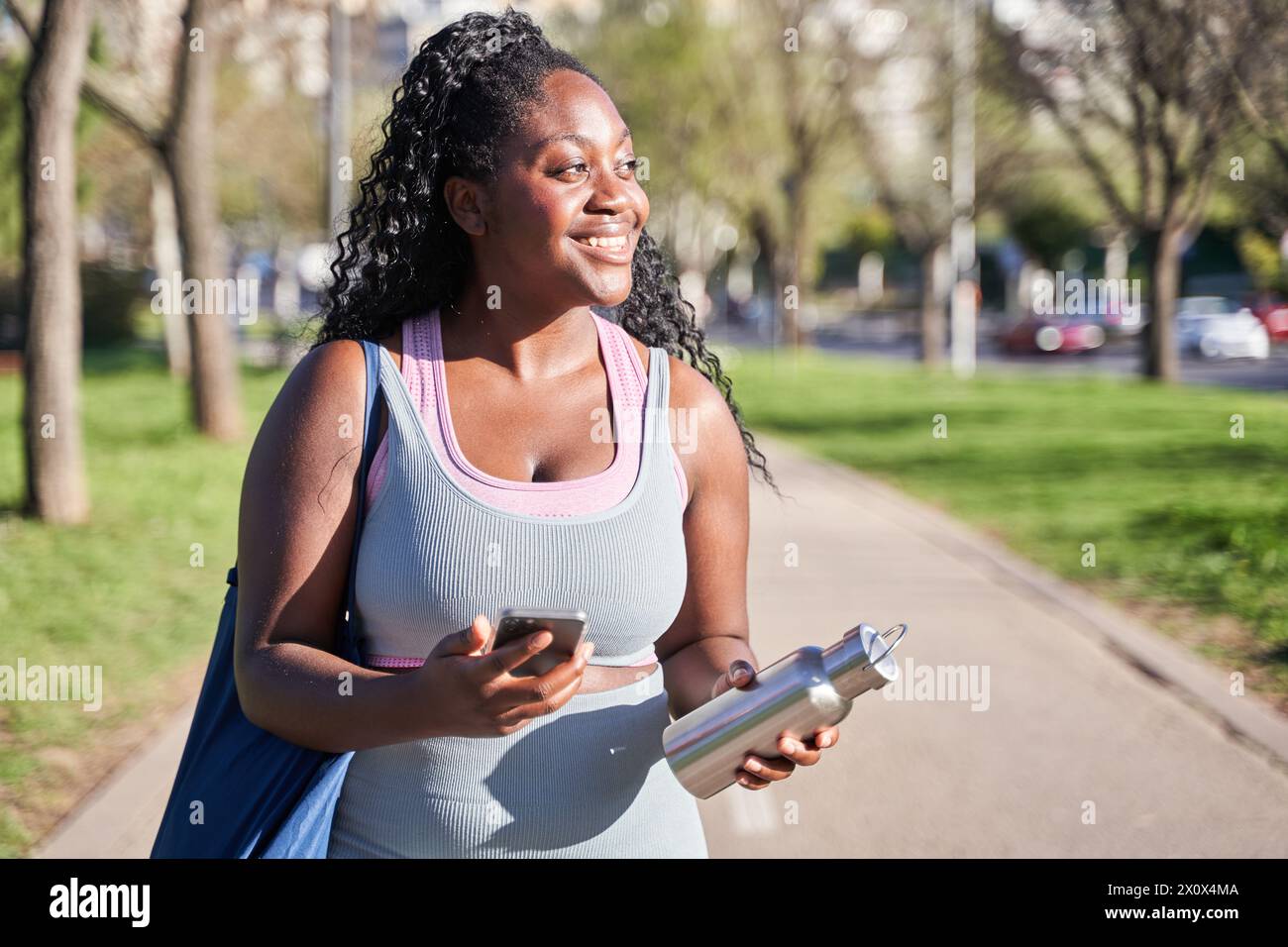 Junge afroamerikanische Frau, die in Sportbekleidung durch den Park geht und ein Smartphone in der einen Hand und eine Flasche Wasser in der anderen hält. Stockfoto