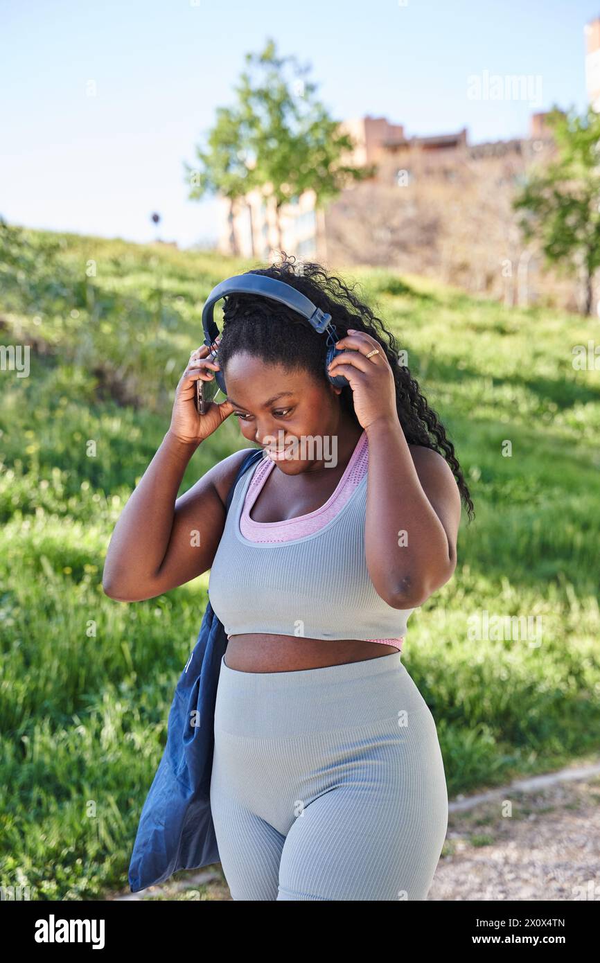 Erfolgreiche kurvige afroamerikanische Frau im Park während des aktiven Trainings und Joggings verwendet Smartphone-App, Sportsfrau lächelt in Kopfhörer und hört zu Stockfoto