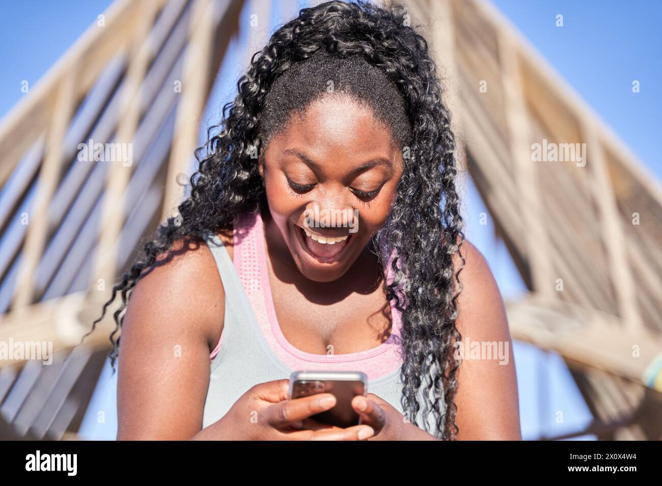 Porträt einer afroamerikanischen Frau mit lockigem Haar, die mit einem überraschten Gesicht auf ihr Smartphone schaut. Stockfoto
