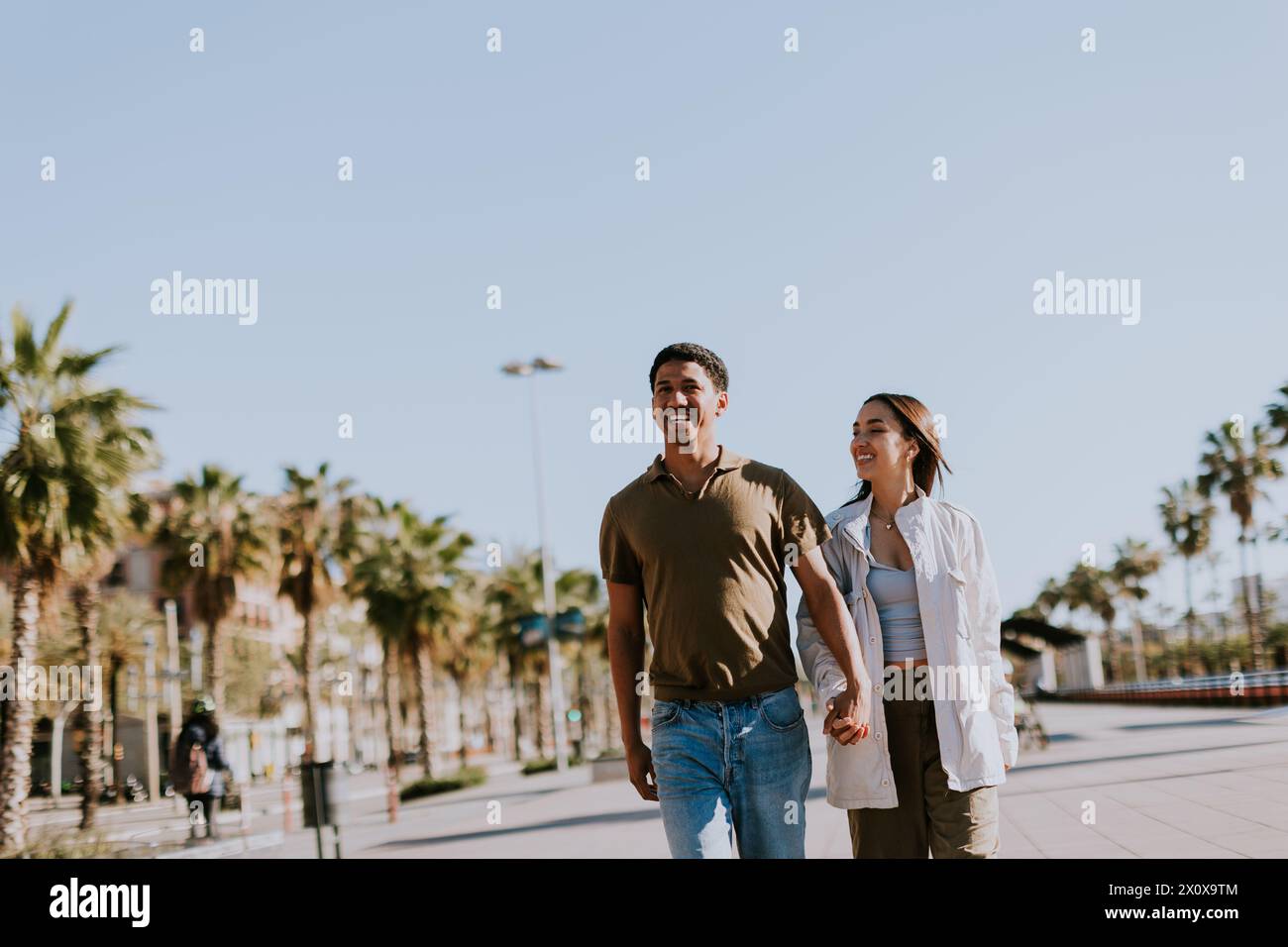 Das junge Paar hält Hände und lächelt, während es einen sonnigen Pfad in Barcelona entlang geht, der von Palmen eingerahmt wird Stockfoto