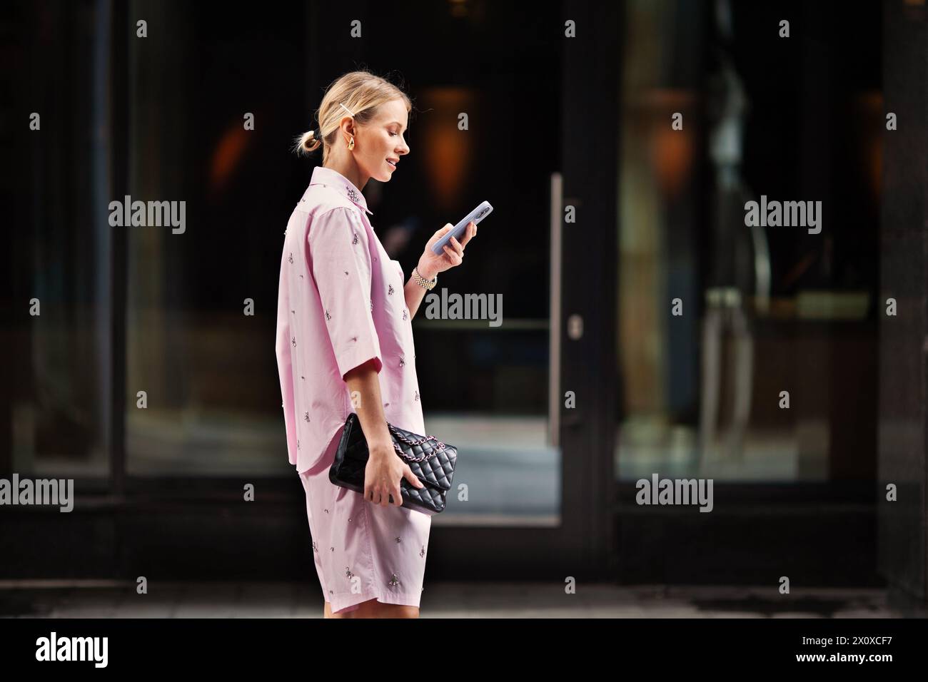 Junge, trendige Frau, die auf ihrem Handy SMS schreibt, fasziniert vom mobilen Bildschirm, während sie auf einer Stadtstraße steht. Städtische Vernetzung und moderner Lifestyle. Stockfoto